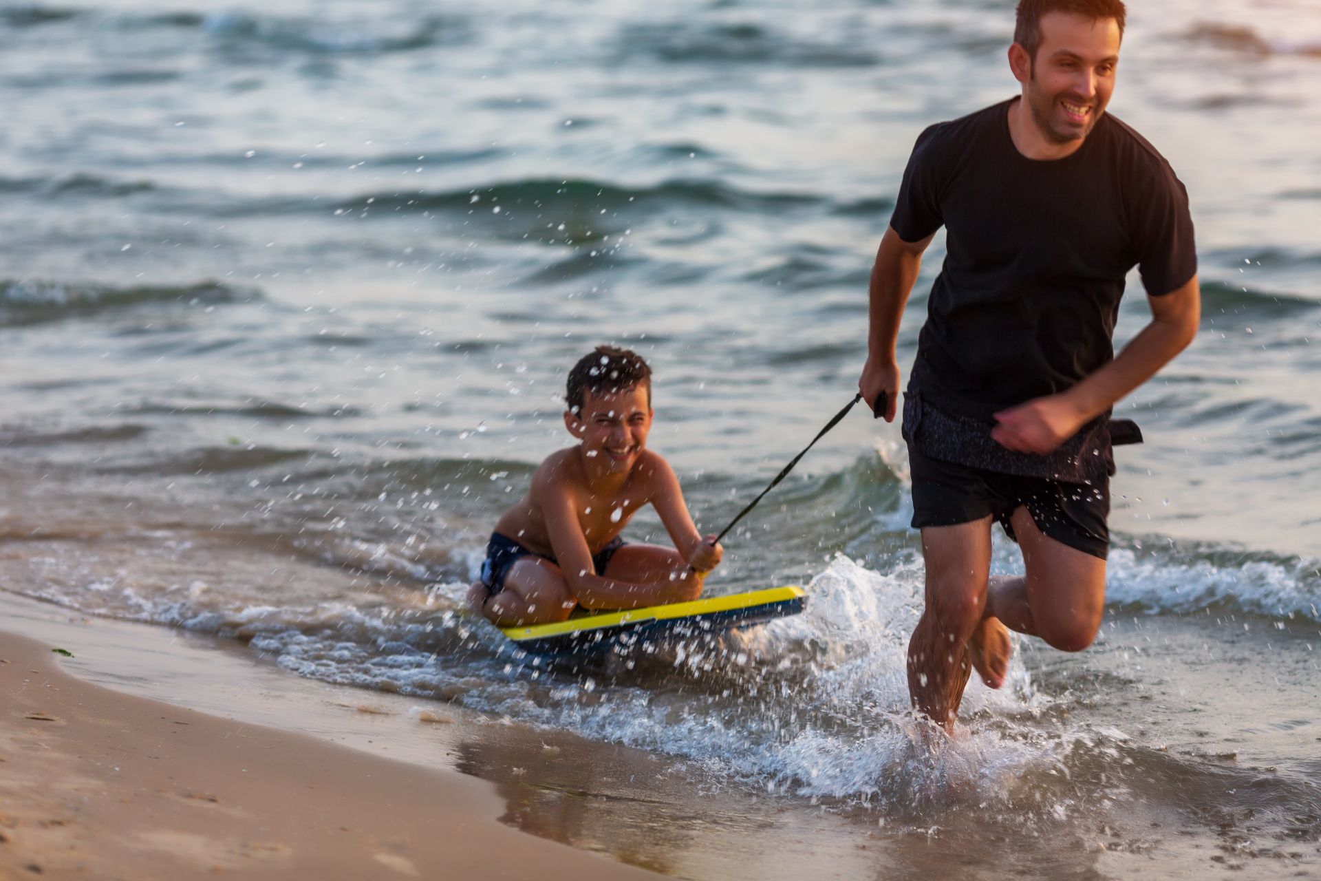 A young boy being pulled on a paddleboard in the waters on a beach by an older boy. He is laughing and enjoying himself